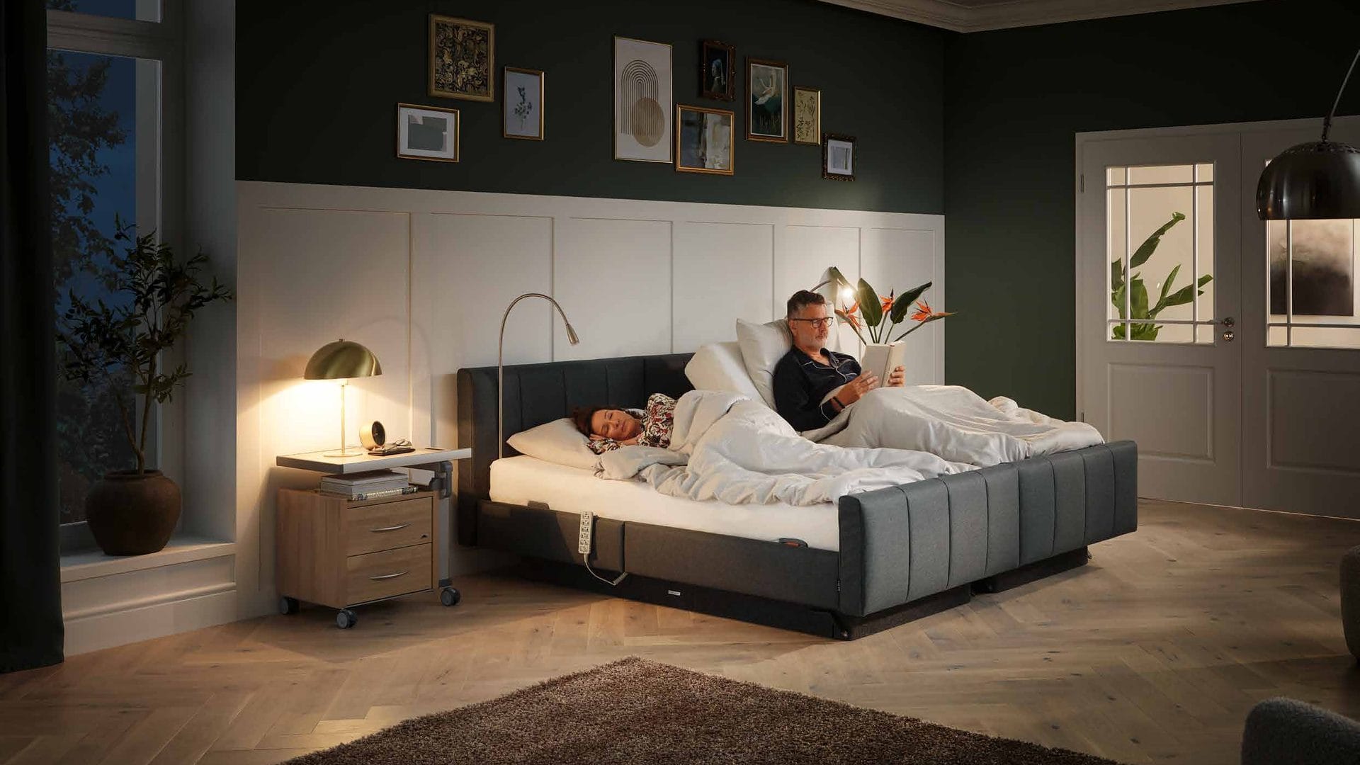Stiegelmeyer Libra deluxe Komfortbett in Anthrazit, abends im Schlafzimmer: Mann liest in aufgerichteter Rückenlehne, Partnerin schläft, Aparto-Nachttisch daneben